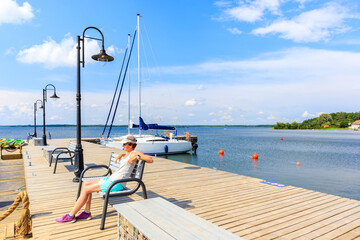 Young woman sitting on pier and looking at boats anchoring in  sailing port Nowe Guty on Lake Sniardwy on summer sunny day,  Masurian Lakes, Poland
