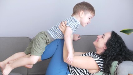 Happy healthy family young mom hugs cute little child son who kisses mother on lips or cheek sitting on sofa at home, funny mother and son kid boy love.