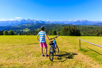 Fototapeta premium Young female biker standing with bicycle on meadow and looking at panorama of Tatra Mountains on beautiful summer sunny day at Lapszanka Pass, Poland