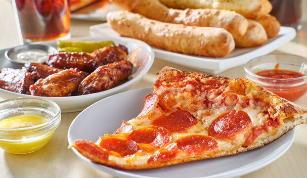 sliced of pepperoni pizza on plate with breadsticks, soft drink and chicken wings in background