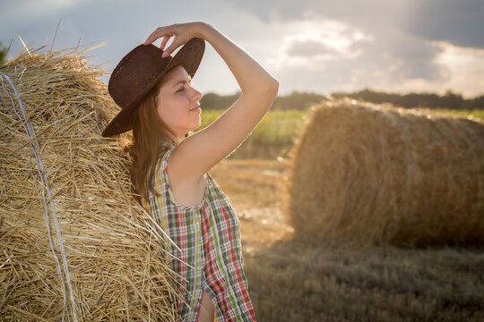 Young girl in a cowboy hat on hay bale. Girl farmer in denim shorts and plaid shirt, in a wheat field in sunset. Happy Woman cowgirl on a farm. Freedom lifestyle.