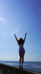 Obraz premium A slender curly-haired girl in denim shorts on the background of the blue sea on a sunny day, summer vacation. model shows V sign, world at peace