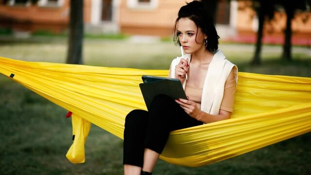 Art work of young woman drawing at tablet computer. Girl relaxes sitting in a hammock in a city park. Student creating image by stylus at display of digital notepad