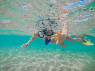 Underwater picture of boy in mask and flippers with blue water and sand coastline. Summertime activity. Snorkeling to see underwater life. Holidays for kids in the sea vacation in tropical country. 
