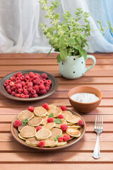Pancake cereals and red ripe fresh raspberries in a brown plate. A bunch of Melissa in a mug on a dark wooden table near the window. Delicious Breakfast