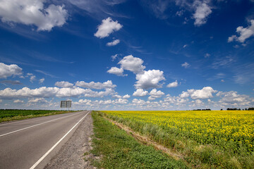 Fototapeta na wymiar Asphalt road through yellow field sunflower and green field corn. Blue Sky and white clouds panoramic view. Beautiful scenic dynamic Landscape agricultural land. Beauty nature, Agriculture. Empty path