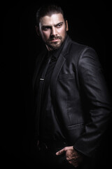 Obraz premium Young white Caucasian man on a black background in a black jacket, shirt and black tie looks out from under his forehead. Strict male portrait. The guy in the classic black suit.