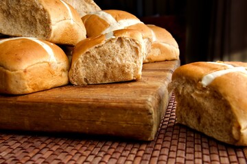 Hot cross buns on a wooden board against a black background.  One bread rolls sits to the side of the wooden board.