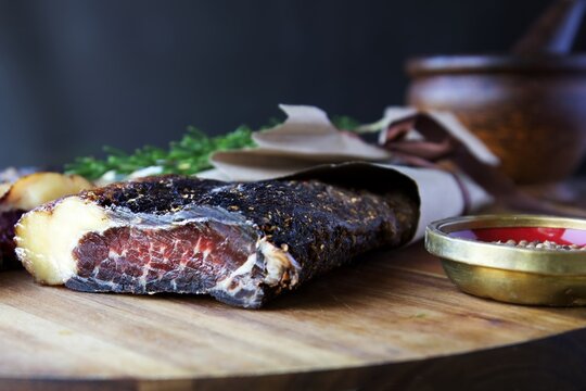 A piece of dried meat biltong on a wooden board wrapped in brown paper.  A small bowl of coriander seed sits to the side and a pestal and mortar against a black background 