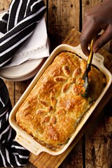 Cooked pie on a wooden surface about to be served up by a black person.  A pile of plates and a black dish towel sits to the side of the dish