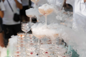 The waiter pours champagne into crystal glasses with dry ice and white smoke close up. Sparkling wine pouring in wine glass at summer day. Catering for business people.