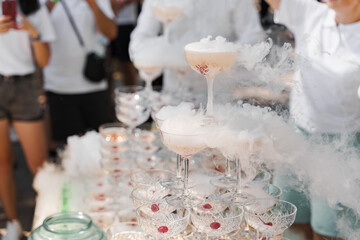 The waiter pours sparkling wine into crystal glasses with dry ice and white smoke close up. Champagne pouring in wine glass at summer day. Catering for wedding ceremony.