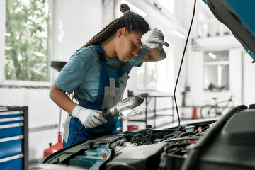 Excellence in performance. Young african american woman, professional female mechanic wiping forehead, examining under hood of car with torch at auto repair shop