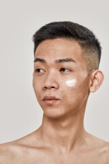 Obraz premium Perfect skincare. Close up portrait of shirtless young asian man with cream applied on his cheek looking aside isolated over white background. Beauty, skincare routine concept
