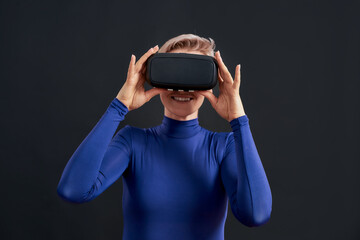 Excited woman with short hair in blue turtleneck wearing virtual reality headset, vr glasses isolated over dark background