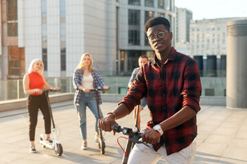 afro american guy with an electric scooter in the company of friends in the city, interracial people use eco transport