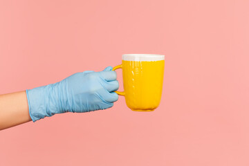 Profile side view closeup of human hand in blue surgical gloves holding and showing yellow cup of hot drink in hand. indoor, studio shot, isolated on pink background.