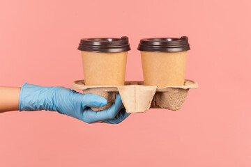 Profile side view closeup of human hand in blue surgical gloves holding and showing cups of hot takeaway mug drink in hand. indoor, studio shot, isolated on pink background.