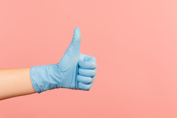 Profile side view closeup of human hand in blue surgical gloves showing like or thumbs up. indoor, studio shot, isolated on pink background.