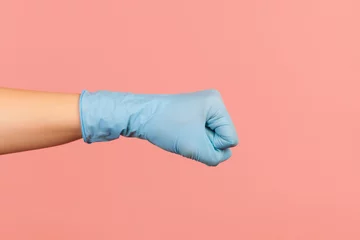 Foto op Plexiglas Profile side view closeup of human hand in blue surgical gloves showing boxing fists with hand  attack or defense against problems. indoor  studio shot  isolated on pink background © khosrork