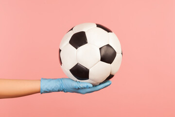 Profile side view closeup of human hand in blue surgical gloves holding football or soccer ball. indoor, studio shot, isolated on pink background.