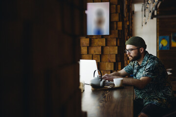 Stylish young bearded male hipster student in eyeglasses studying in cafe.