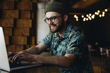 Smiling young smart bearded male hipster in eyeglasses and hat having video meeting on laptop in online communications platform