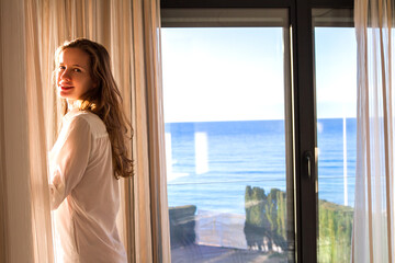 The beautiful woman standing near balcony window indoor in the morning at sunrise.