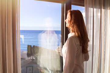 Beautiful long-haired woman in a white shirt standing indoor by a panoramic window with curtains...