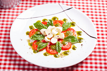 Traditional Italian caprese salad with sliced tomatoes, mozzarella cheese, basil and pesto sauce on white plate. Close up
