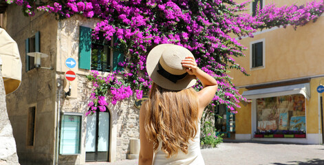 Back view of young tourist woman holding hat visiting Sirmione old flowered town on Lake Garda, Italy