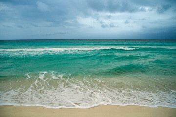 Tropical landscape. Seascape. The beach under a dramatic cloudy sky. The white sand shore, turquoise color water ocean, sea waves and horizon.