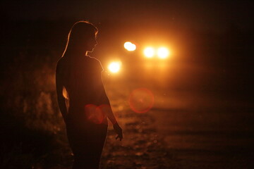 Silhouette of young slender woman in the backlight of car headlights on the road
