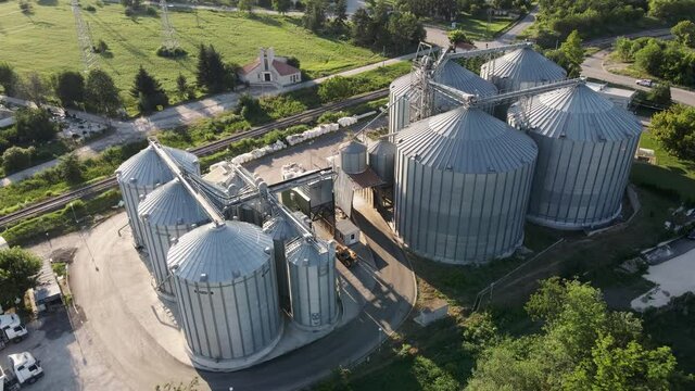 Warehouse granary  aluminum containers for storing loose grain against blue sky and voluminous clouds. Elevator. Parked trucks. Large granary in field on background of sunset aerial view drone turn