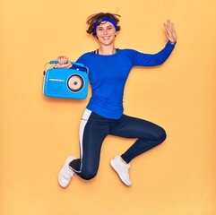Young beautiful sportswoman listening to music smiling happy. Jumping with smile on face holding vintage radio over isolated yellow background.