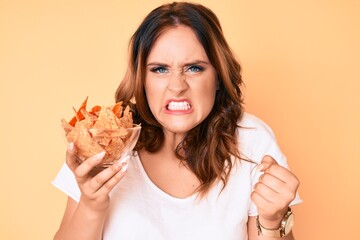 Young beautiful caucasian woman holding nachos potato chips annoyed and frustrated shouting with...