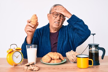 Senior handsome man with gray hair sitting on the table eating croissant for breakfast stressed and frustrated with hand on head, surprised and angry face