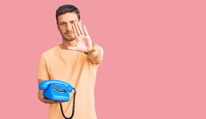 Handsome young man with bear holding vintage telephone with open hand doing stop sign with serious and confident expression, defense gesture