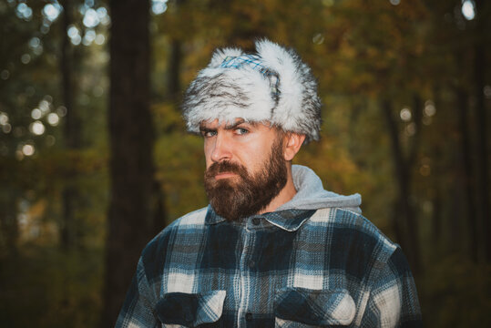 Bearded man in winter fur cap with ear-flaps eye askance at camera. Stylish male wearing plaid shirt and ear-flaps fur cap at autumn fall trees background. Cold autumn weather concept.