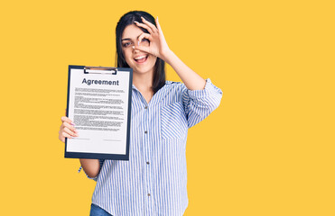 Young beautiful girl holding clipboard with agreement document smiling happy doing ok sign with hand on eye looking through fingers