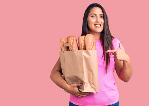 Young beautiful brunette woman holding delivery bag with bread smiling happy pointing with hand and finger