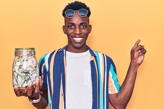 Young african american man holding jar with savings smiling happy pointing with hand and finger to the side