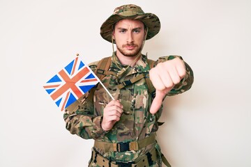 Fototapeta premium Young caucasian man wearing camouflage army uniform holding united kingdom flag with angry face, negative sign showing dislike with thumbs down, rejection concept