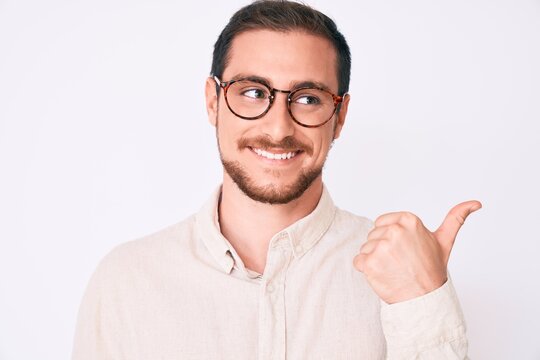 Young handsome man wearing casual clothes and glasses smiling with happy face looking and pointing to the side with thumb up.