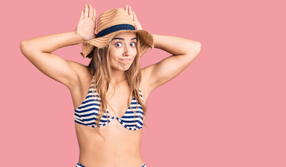 Young beautiful blonde woman wearing bikini and hat doing bunny ears gesture with hands palms looking cynical and skeptical. easter rabbit concept.