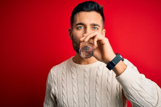 Young handsome man drinking glass of healthy water to refreshment standing over isolated red background