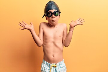 Cute blond kid wearing swimwear and swimmer glasses clueless and confused with open arms, no idea and doubtful face.