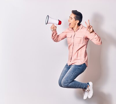 Young beautiful caucasian woman screaming using megaphone. Jumping doing victory sign over isolated white background