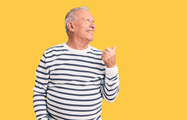 Senior handsome grey-haired man wearing casual striped sweater smiling with happy face looking and pointing to the side with thumb up.