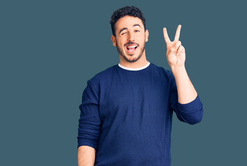 Fototapeta premium Young hispanic man wearing casual clothes smiling with happy face winking at the camera doing victory sign. number two.